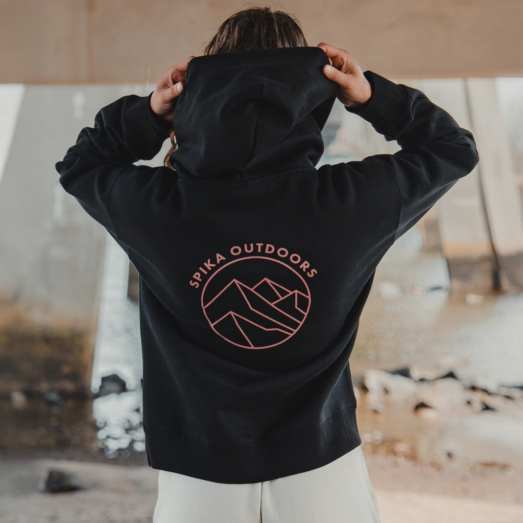 Woman pulling up the hood of the Spika GO Contour black hoodie, showing the mountain back print