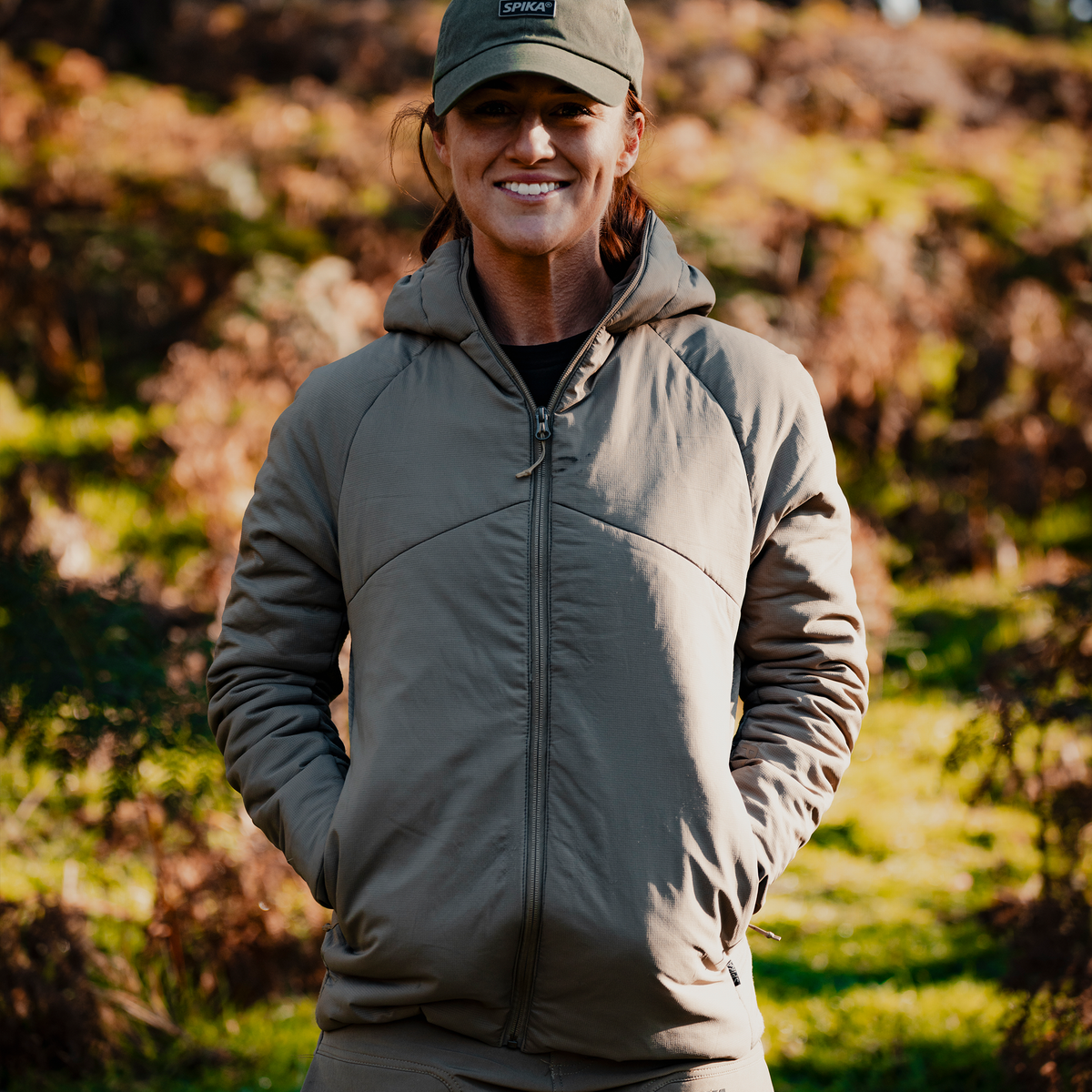 Woman wearing the Spika Aura hybrid puffer jacket in Dusky Green and a cap, smiling outdoors in bushland