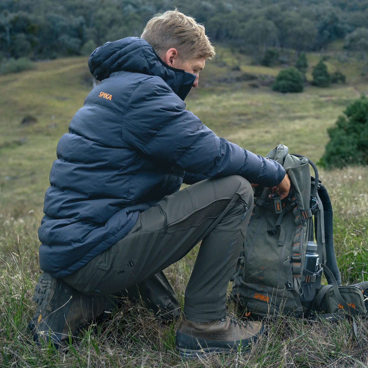 Hunter kneeling in grassland beside a backpack wearing a Spika Terra men's puffer jacket