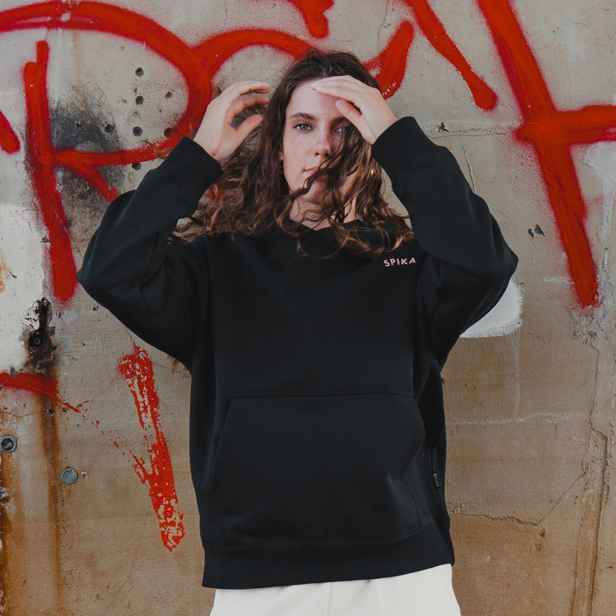 Woman wearing the Spika GO Contour women's black hoodie, hands in her hair against a graffiti wall