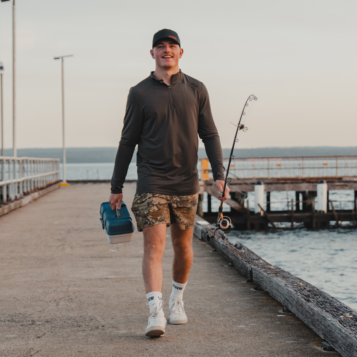 Man walking along a jetty with a tackle box and rod wearing a Spika Enduro quarter-zip top