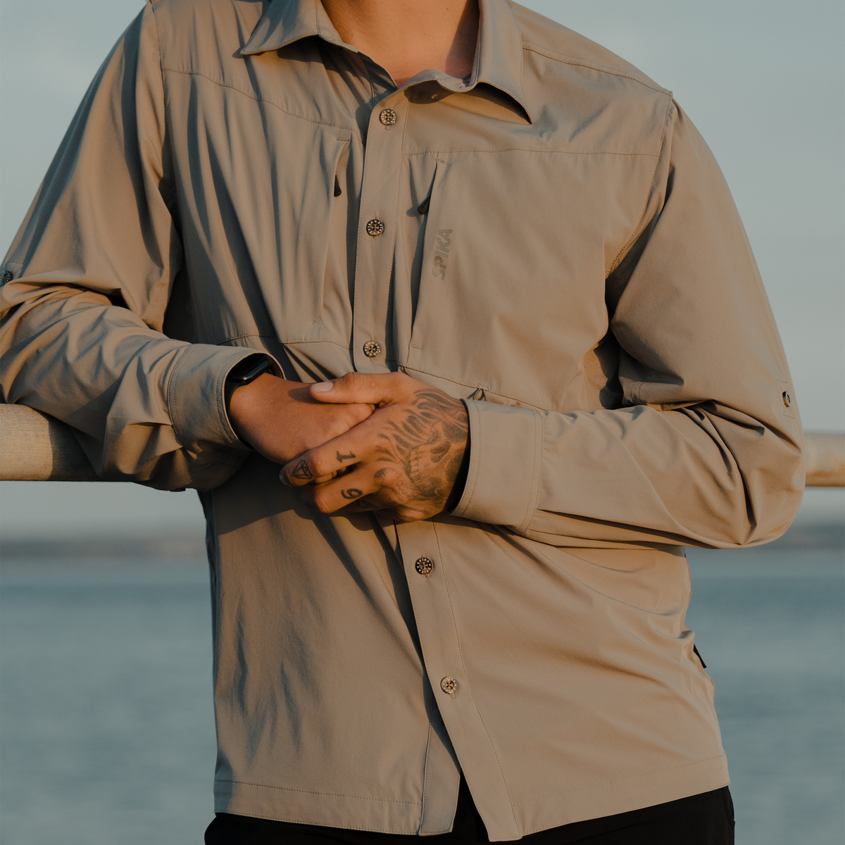 Man wearing Spika Skipper men's shirt in Marine, leaning on a waterfront railing at sunset