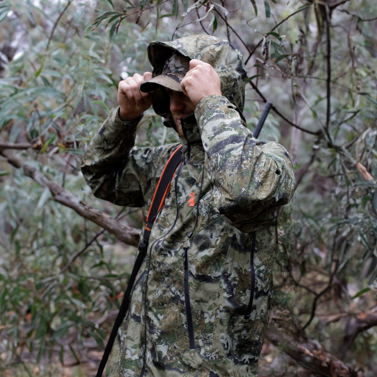 Hunter pulling up the hood of his Spika Edge Biarri Camo jacket in wet bushland, rifle slung