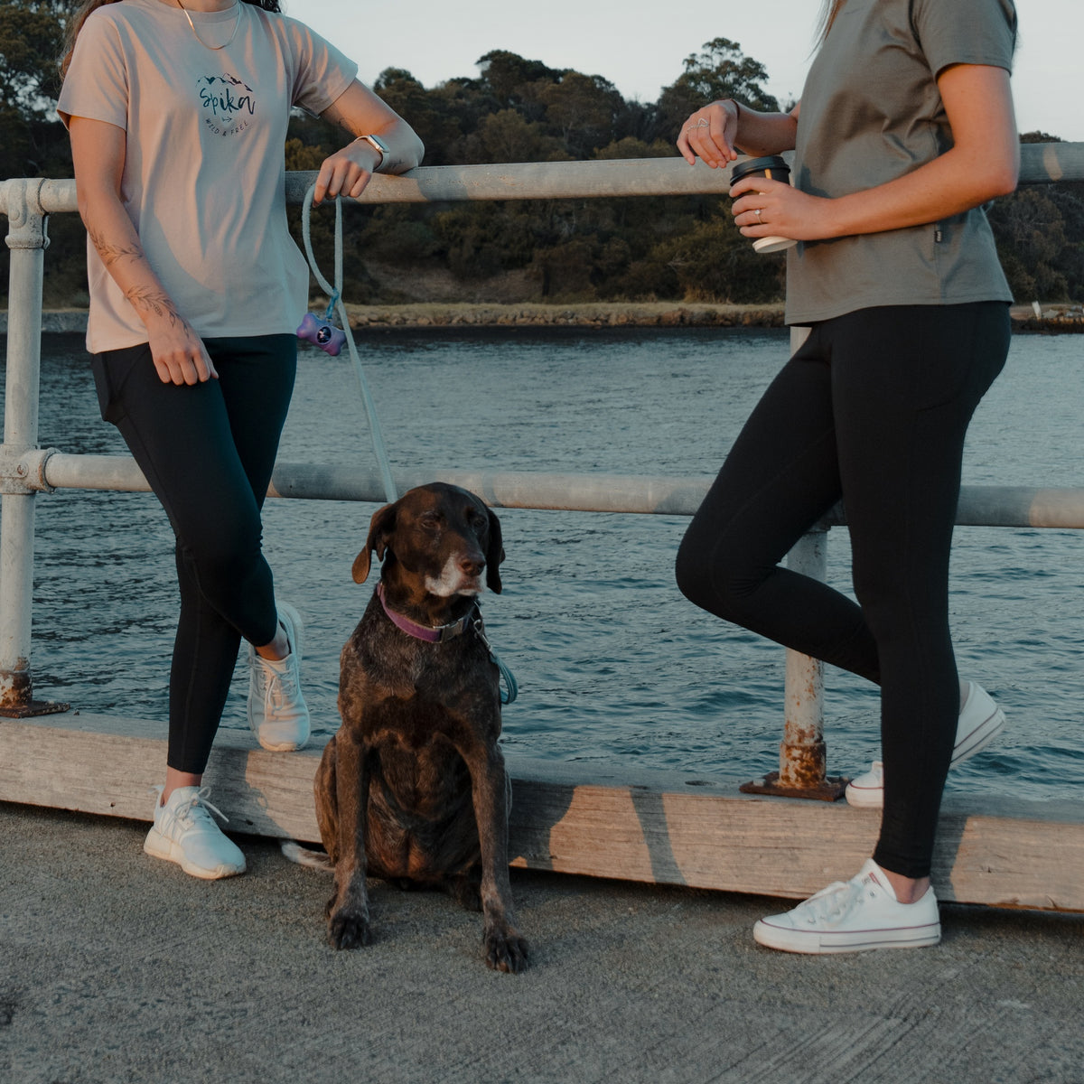 Two women with a dog leaning on a jetty railing, both wearing Spika GO Ember black leggings