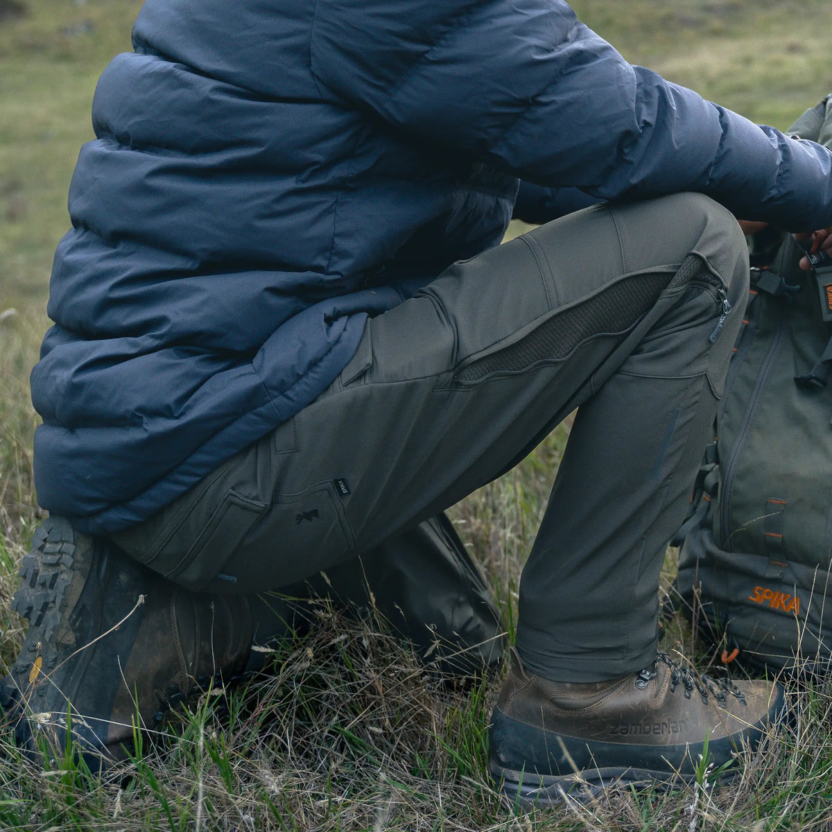 Hunter crouching in grass wearing Spika Recon men's pants with a puffer jacket and hiking boots