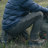 Hunter crouching in grass wearing Spika Recon men's pants with a puffer jacket and hiking boots