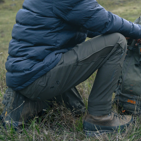 Hunter crouching in grass wearing Spika Recon men's pants with a puffer jacket and hiking boots