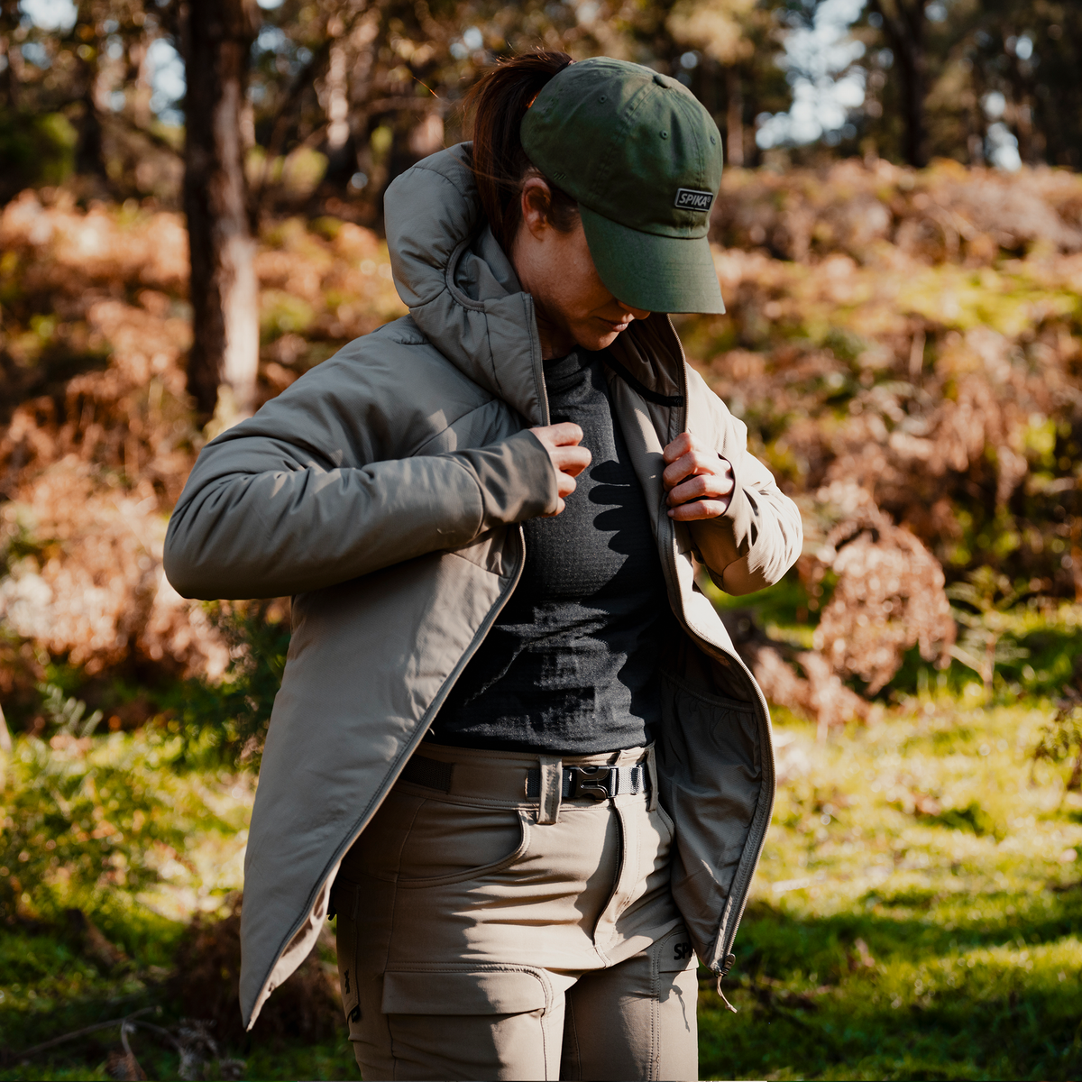 Woman zipping up the Spika Aura hybrid puffer jacket in Dusky Green while standing in sunlit bush