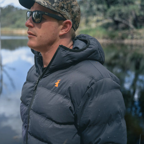 Man in a camo cap wearing the Spika Terra men's puffer jacket in Black beside a river