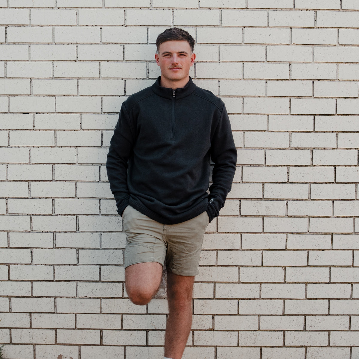 Man wearing the Spika GO Belay men's slate quarter-zip jumper with tan shorts against a brick wall