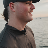 Smiling man by the water in a cap and the Spika Enduro Olive Summit athletic top, close-up