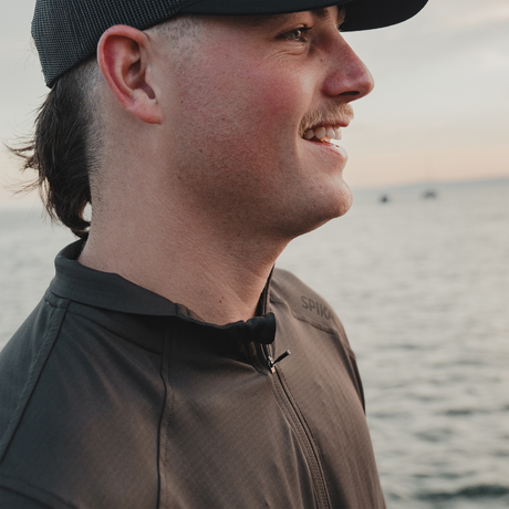 Smiling man by the water in a cap and the Spika Enduro Olive Summit athletic top, close-up
