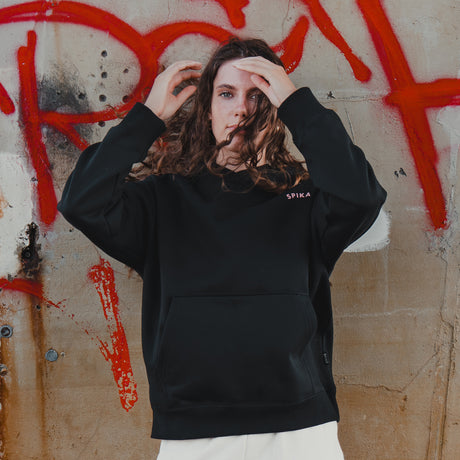 Woman wearing the Spika GO Contour women's black hoodie, hands in her hair against a graffiti wall