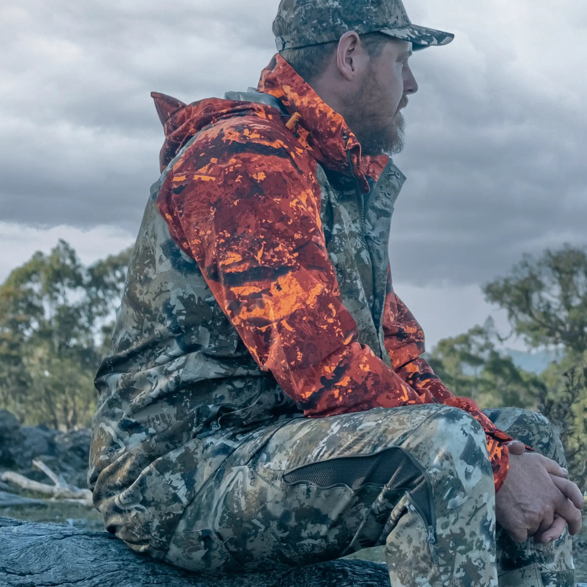 Hunter in the Spika Valley men's jacket in Biarri Camo/Blaze sitting on a rock outdoors
