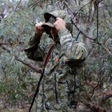 Hunter adjusting the hood of a Spika Edge waterproof jacket among eucalypts, rifle slung