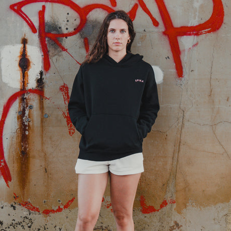 Woman in the Spika GO Contour black hoodie and cream shorts standing at a graffiti wall, full length