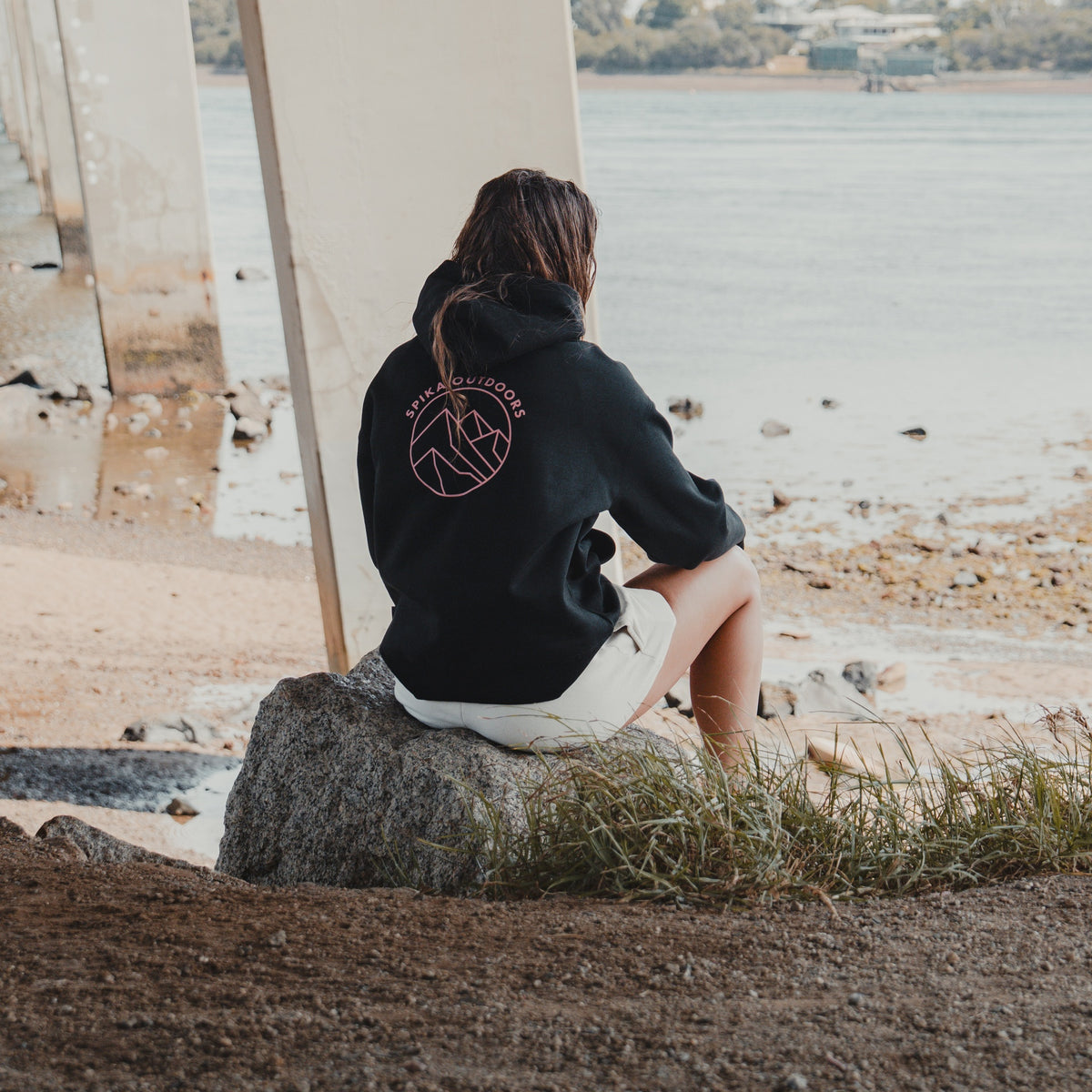 Woman in the Spika GO Contour black hoodie sitting on a rock by the water, showing the back print