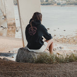 Woman in the Spika GO Contour black hoodie sitting on a rock by the water, showing the back print