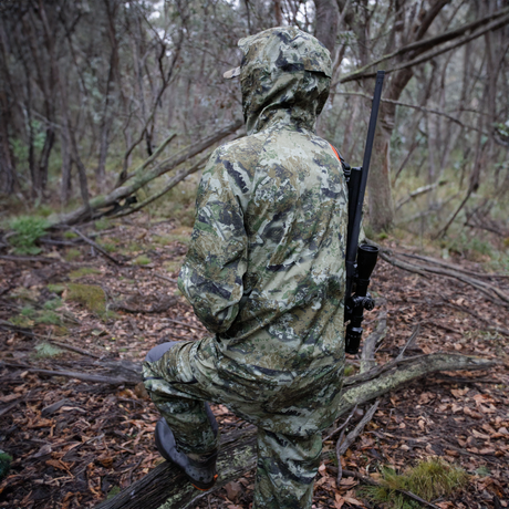 Hunter in the Spika Edge Biarri Camo jacket kneeling on a log with a rifle slung on his back