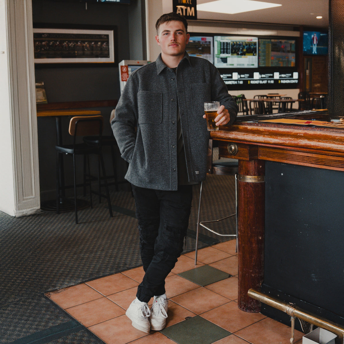 Man wearing the Spika GO Civvy slate shirt jacket with black pants, leaning on a pub bar with a beer