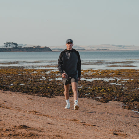 Man in a cap walking along the beach wearing the Spika GO Scuba men's slate and black hoodie