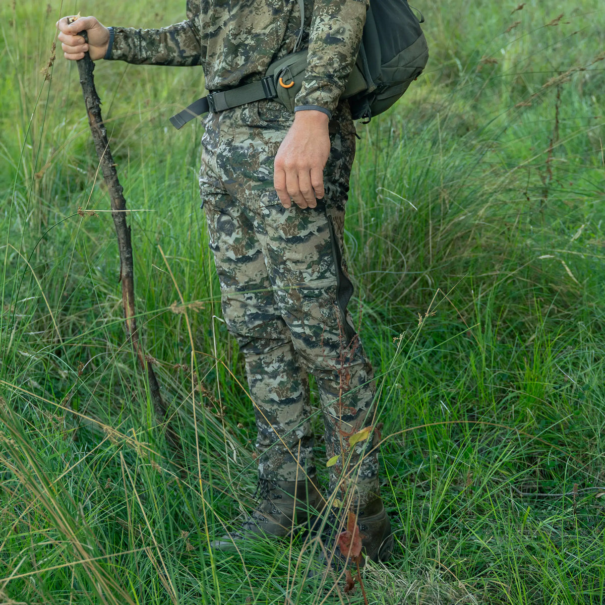 Hunter wearing Spika Recon men's pants in Biarri Camo standing in long grass with a walking stick