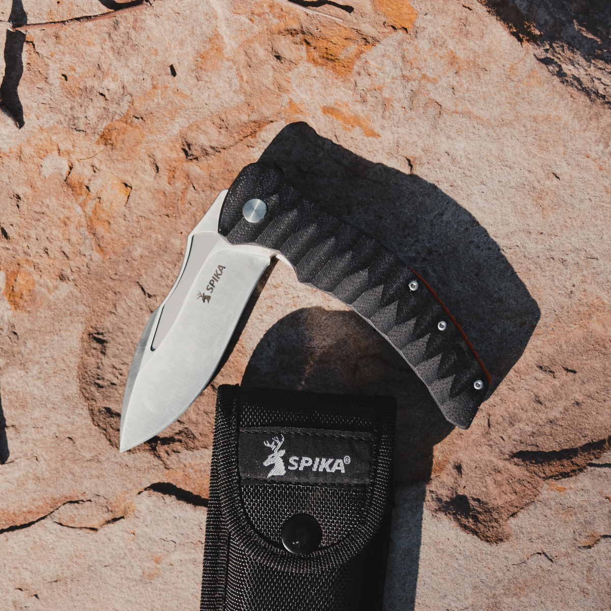 Spika Bushmaster Viper folding knife lying on red rock beside its black nylon Spika belt pouch