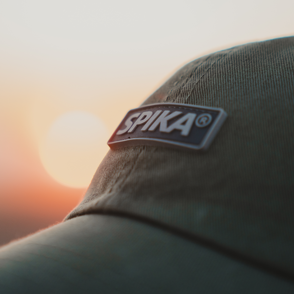 Spika GO Advance dad cap in Olive, close-up of the Spika logo patch against a sunset