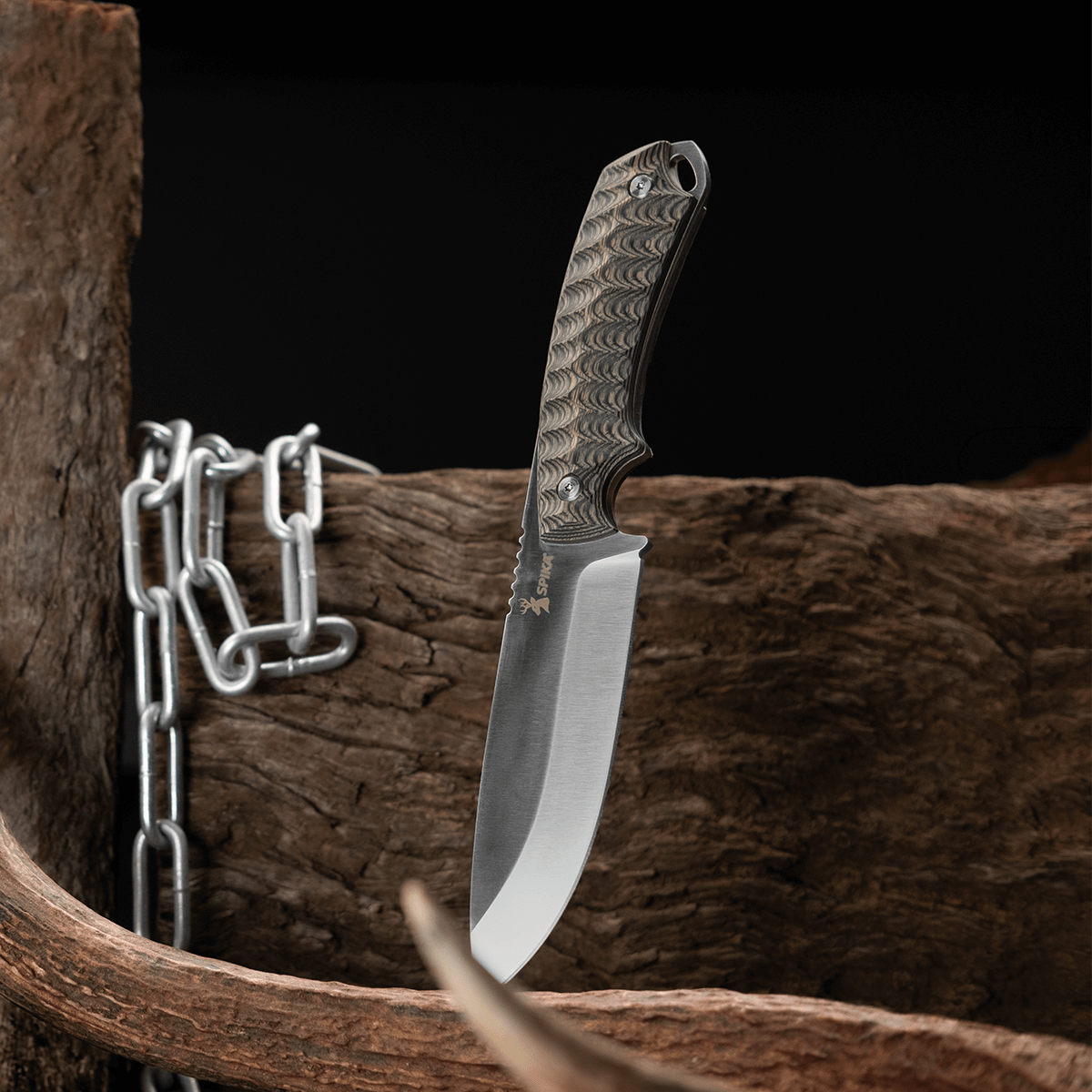 Spika Bushmaster Dogtooth knife standing blade down beside timber, antler and a steel chain