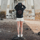 Woman with the hood up on the Spika GO Contour black hoodie, rear view on a rocky shoreline