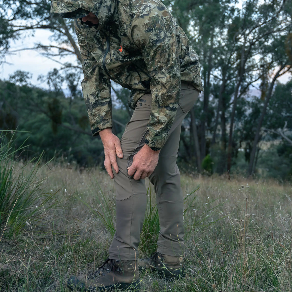Hunter in camo jacket wearing Spika Recon men's pants, bending to adjust the knee in a grassy clearing
