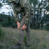 Hunter in camo jacket wearing Spika Recon men's pants, bending to adjust the knee in a grassy clearing
