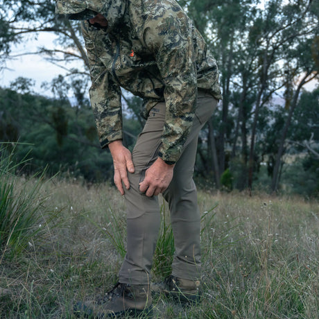 Hunter in camo jacket wearing Spika Recon men's pants, bending to adjust the knee in a grassy clearing